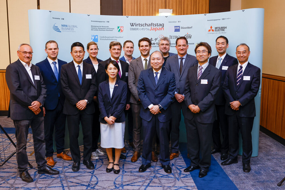 Fourteen people in business attire stand in a group, with a screen displaying logos and lettering in the background.