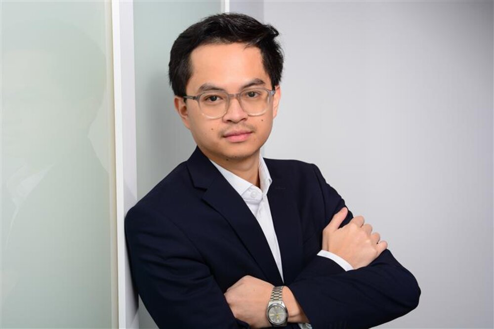 A portrait photograph of a smartly dressed young man wearing glasses and a watch. He is leaning with one shoulder against an office wall.