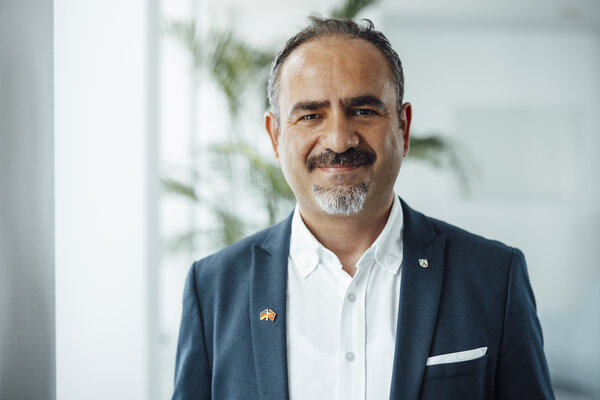 A portrait photograph of a smartly dressed man with a small beard in an office.
