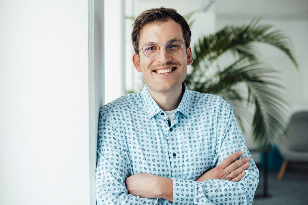A portrait photograph of a man leaning sideways against an office wall. He is wearing glasses and a light-coloured patterned shirt. 