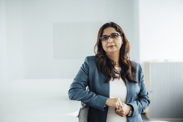 A portrait photograph of a smartly dressed woman standing next to an office desk and leaning on it.