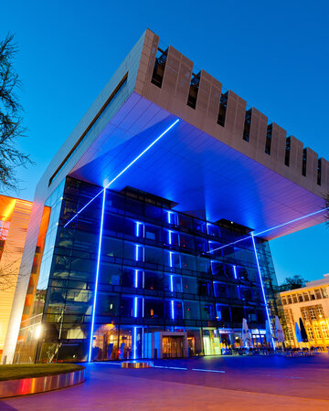 [Translate to 中文:] Modern building with blue lighting and glass façade at night, surrounded by trees and street lights.