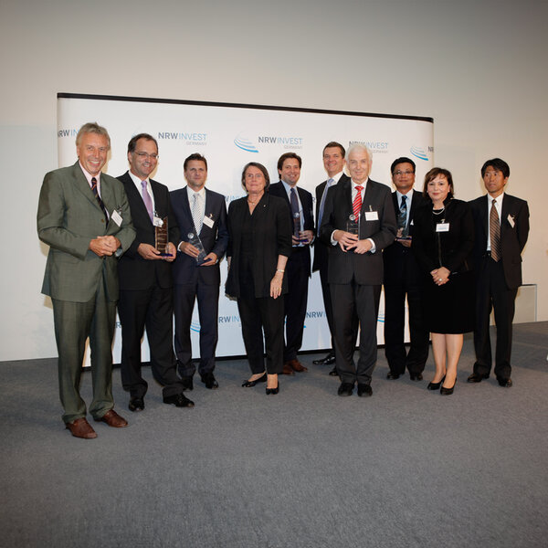 Ten people in business attire stand side by side, several holding glass trophies, behind them a screen with the lettering NRW Invest Germany.