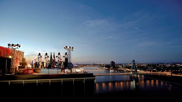 An open-air concert on a rooftop at sunset with views over a big city and a river with illuminated bridges.