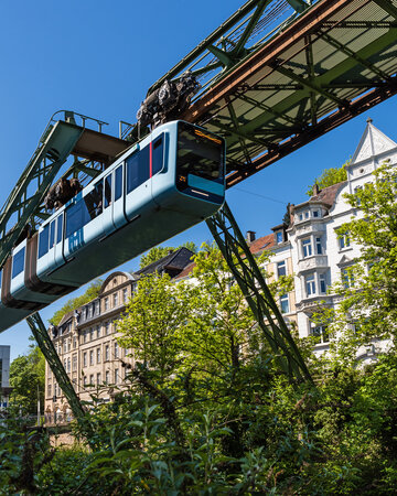 [Translate to 中文:] Wuppertal Suspension Railway running on an elevated track between trees and buildings in the Bergisches Städtedreieck region.