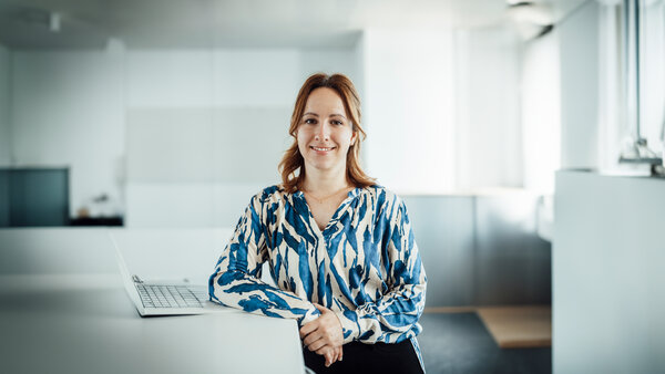 A portrait photo of a young woman. She is wearing a patterned blue and white blouse. She is standing at an office desk with a laptop on it.