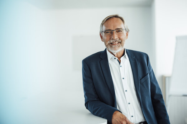 A portrait photo of a smartly dressed man standing sideways at an office desk with his arm resting on it. He is wearing glasses.