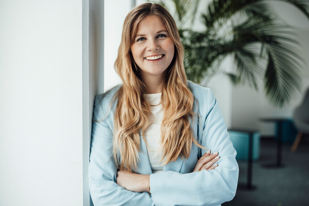 A portrait photograph of a young woman on an office wall. She is wearing a light blue blazer and smiling at the camera.