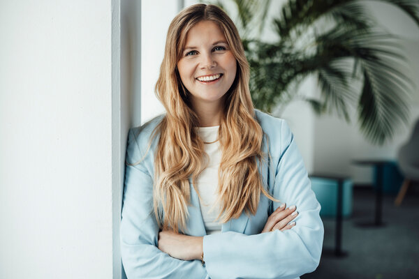 A portrait photograph of a young woman on an office wall. She is wearing a light blue blazer and smiling at the camera.