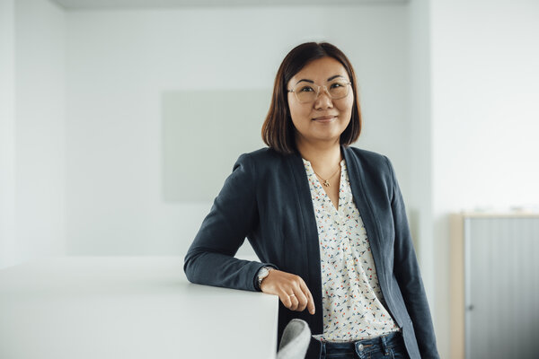 A portrait photograph of a woman standing next to an office desk with one arm resting on it. She is wearing glasses, a necklace and a watch.