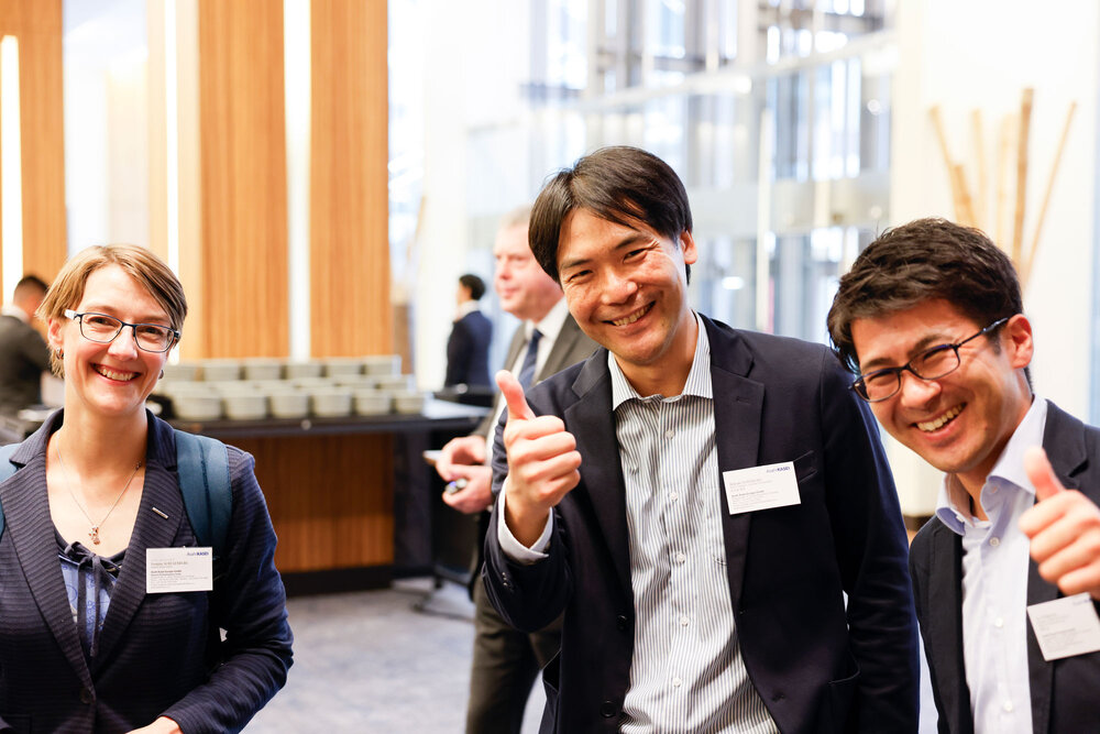 Three people in business attire smile at the camera, two give a thumbs up, blurred background with other people.