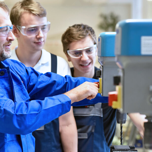 Zwei Auszubildende schauen ihrem Ausbilder bei der Bedienung einer Bohrmaschine zu.