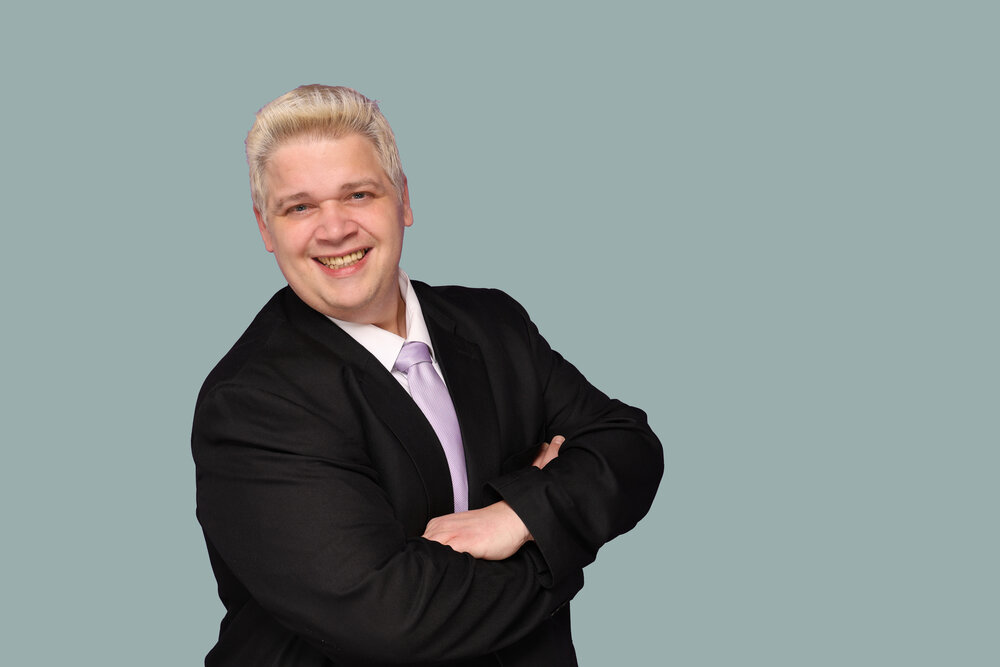 A portrait photograph of a smartly dressed man standing sideways to the camera with his arms crossed and smiling.