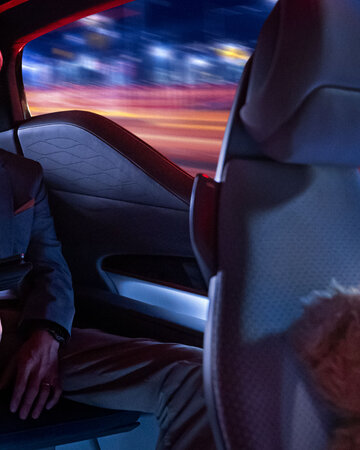 Ein Mann sitzt auf dem Rücksitz eines Autos mit Tablet in der Hand, neben ihm liegt ein Teddybär auf dem Vordersitz, draußen ziehen nachts Lichter vorbei.