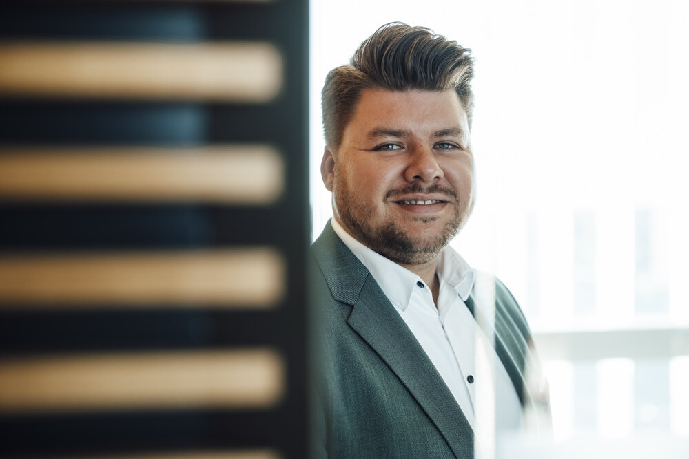 A portrait photograph of a young man with styled dark blond hair, wearing a white shirt and a grey jacket.