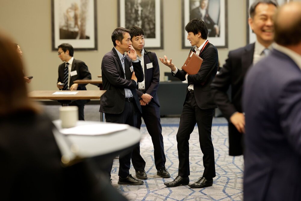Several men in business attire are standing in a group and conversing, one is holding a brown folder, blurred foreground with additional people.