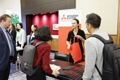 Several people at an information stand, a woman holding a red bag, a Mitsubishi Electric banner in the background.