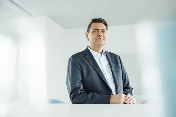 A portrait photo of a man standing behind an office desk with his hands resting on it. He is wearing a light-coloured shirt and a dark jacket.