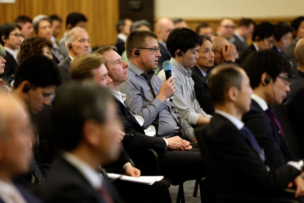Several people in business attire are seated in the audience, and a man wearing glasses and a headset is speaking into a microphone.