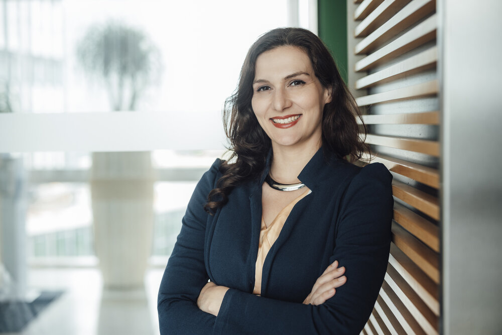 A portrait photograph of a smartly dressed woman with her arms crossed and jewellery around her neck in front of a large window.