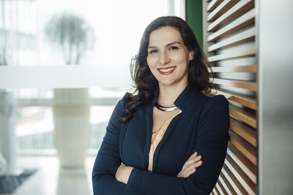 A portrait photograph of a smartly dressed woman with her arms crossed and jewellery around her neck in front of a large window.